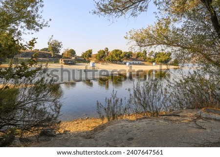 a beautiful summer landscape at Puddingstone Lake with a sandy beach, a lifeguard tower, lush green trees, rippling water, blue sky and clouds at sunset in San Dimas California USA Royalty-Free Stock Photo #2407647561