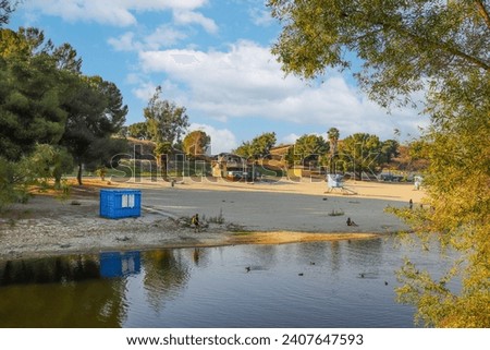 a beautiful summer landscape at Puddingstone Lake with a sandy beach, a lifeguard tower, lush green trees, rippling water, blue sky and clouds at sunset in San Dimas California USA Royalty-Free Stock Photo #2407647593
