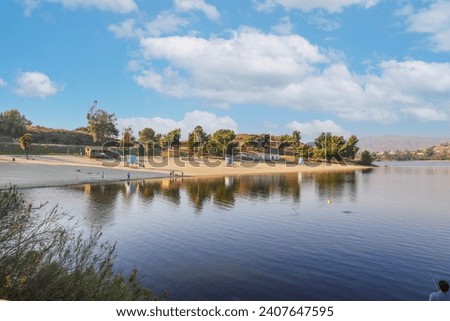 a beautiful summer landscape at Puddingstone Lake with a sandy beach, a lifeguard tower, lush green trees, rippling water, blue sky and clouds at sunset in San Dimas California USA Royalty-Free Stock Photo #2407647595