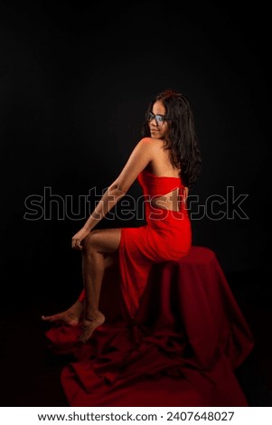 Portrait of beautiful young woman wearing glasses and in red dress sitting posing for photo. Isolated on black background.