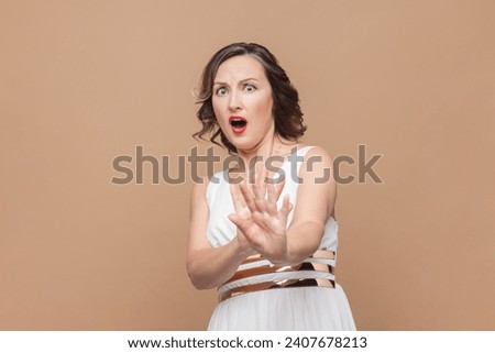 Scared middle aged woman with wavy hair keeps palms forward camera tries to defence himself, feels very frightened, wearing white dress. Indoor studio shot isolated on light brown background. Royalty-Free Stock Photo #2407678213