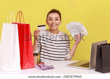 Electronic money. Satisfied happy woman sitting office workplace with laptop, holding dollars and credit card, looking at camera with smile. Indoor studio studio shot isolated on yellow background.