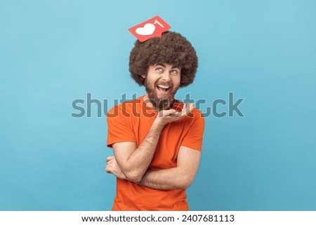 Laughing man with Afro hairstyle wearing orange T-shirt holding like icon above head, looking with funny satisfied facial expression, social media. Indoor studio shot isolated on blue background.