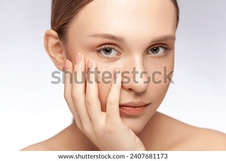 Closeup portrait of serious beautiful woman with natural makeup keeps hand under chin looking at camera with calm expression. Indoor studio shot isolated over gray background.