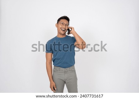 A cheerful, happy Asian man in casual clothes is enjoying talking on the phone with someone, standing on an isolated white studio background. wireless technology, phone calling, communication