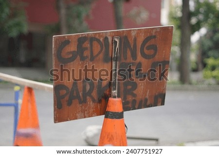 A sign that says "gedung elit parkir sulit" in Indonesian which means it has an elite building but parking is difficult is attached to the road cone