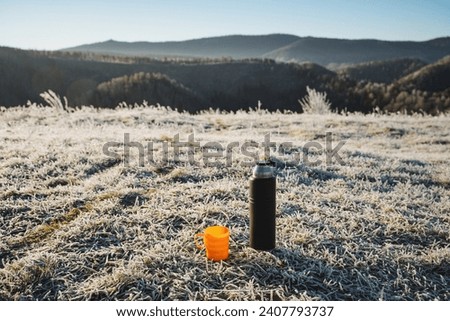 Black thermos stands on the grass against the background of mountains, hot drink in thermal bottle, orange mug, drinking hot tea outdoors in winter. High quality photo