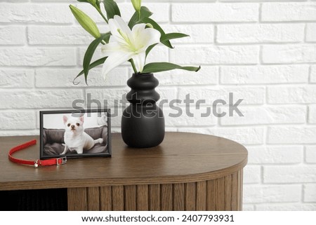 Frame with picture of dog, collar and lily flowers on wooden table near light brick wall. Pet funeral