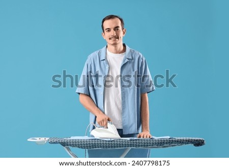 Young man ironing t-shirt on board against blue background