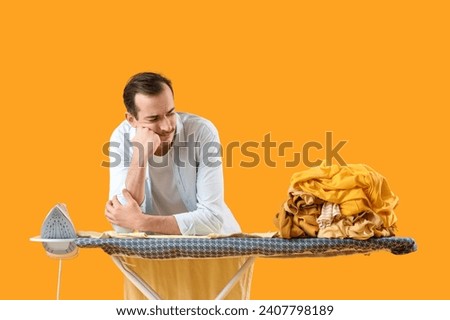 Upset young man with iron and stack of clothes on board against yellow background