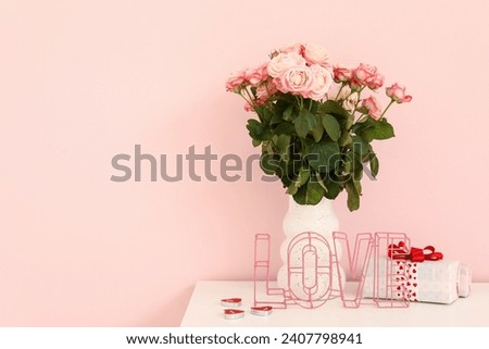 Vase with bouquet of roses and gift box on white cabinet near pink wall, closeup. Valentine's Day celebration