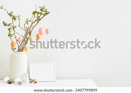 Vase with tree branches, Easter eggs and blank picture frame on table near light wall