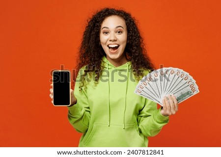 Young amazed woman of African American ethnicity wear green hoody casual clothes hold fan of cash money in dollar banknotes use blank screen mobile cell phone isolated on plain red orange background