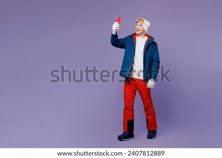 Full body skier man wear warm blue windbreaker jacket ski goggles mask hat doing selfie shot on mobile cell phone spend extreme weekend winter season in mountains isolated on plain purple background