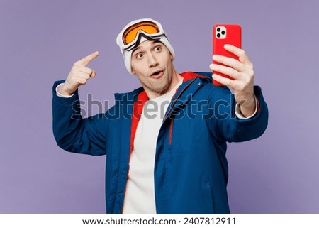 Skier man wear warm blue windbreaker jacket point finger on ski goggles mask hat do selfie shot mobile cell phone spend extreme weekend winter season in mountains isolated on plain purple background