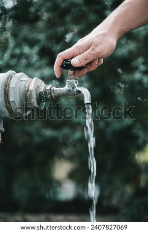 A photograph of a man's hand opening the water tap valve with water flowing out of it. Royalty-Free Stock Photo #2407827069