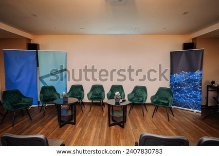 modern meeting room setup with a semi-circle of green velvet chairs around small round tables, each adorned with a plant, and abstract blue graphic roll-up banners in the background. Royalty-Free Stock Photo #2407830783