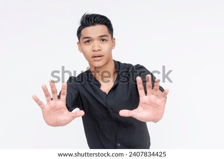 A young asian man puts his hands forward, diffusing a tense situation. Telling someone to calm down. Wearing black polo shirt, isolated on a white background. Royalty-Free Stock Photo #2407834425