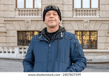 Portrait of a 50-55 year old man in a blue jacket against a blurred background of a city building. Perhaps he is a buyer or an actor, a driver, a loader or a military pensioner. Royalty-Free Stock Photo #2407837599