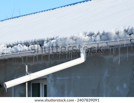 Frozen gutters damage. House  roof covered snow, icicles and frozen roof gutter with downspout pipe. Royalty-Free Stock Photo #2407839091