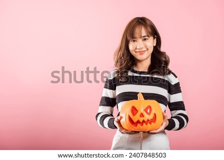 A Halloween-inspired studio shot on pink background features an Asian woman holding model pumpkins, including a ghost-shaped one. Her joyful expression and festive costume enhance the Halloween vibe.