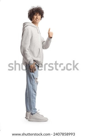 Full length portrait of a tall guy with curly hair in a gray hoodie and jeans gesturing thumbs up isolated on white background