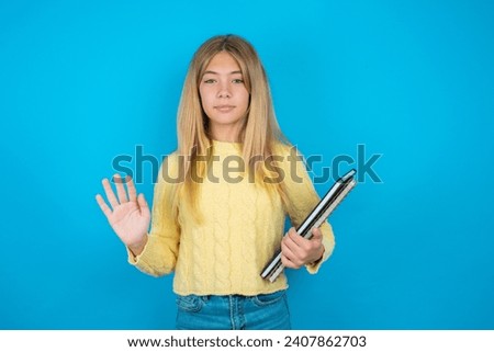 Serious Beautiful kid girl wearing yellow sweater holding notebook pulls palms towards camera, makes stop gesture, asks to control your emotions and not be nervous