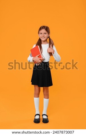 Happy schoolgirl with books showing thumb up on orange background Royalty-Free Stock Photo #2407873007