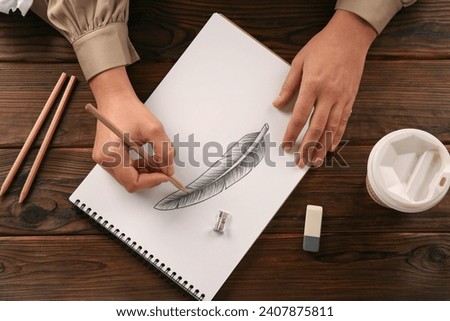 Woman drawing feather with graphite pencil in sketchbook at wooden table, top view