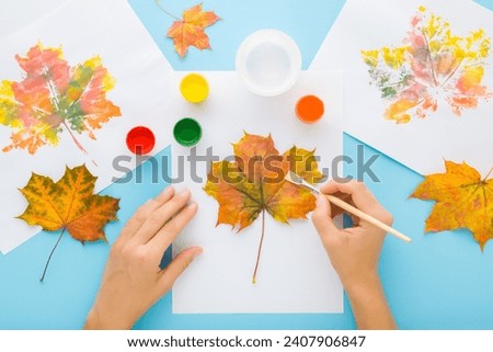 Young adult woman hands painting and copying colorful leaf pattern with gouache color on white paper on light blue table background.  Pastel color. Closeup. Point of view shot.