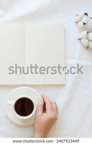 Opened note, cotton branch, hand with a coffee cup on a white sheet. Cozy composition on a bed