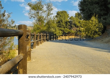 a dirty road to nowhere with a wooden fence along the trail and lush green and autumn colored trees hanging over the path at Puddingstone Lake in San Dimas California USA Royalty-Free Stock Photo #2407942973