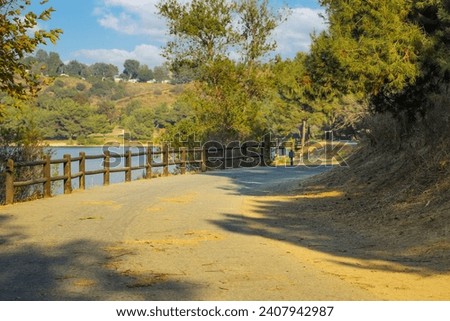 a dirty road to nowhere with a wooden fence along the trail and lush green and autumn colored trees hanging over the path at Puddingstone Lake in San Dimas California USA Royalty-Free Stock Photo #2407942987