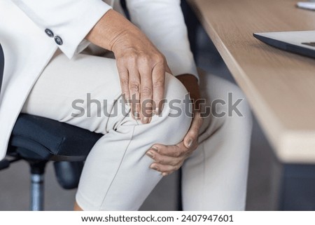 Close-up photo in the office at the desk. The hands of an older woman in a business suit are holding the knee of the leg, giving a massage.