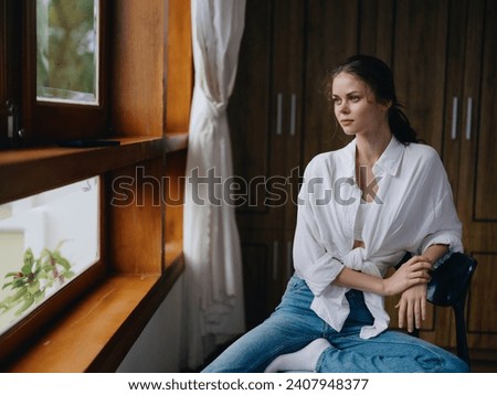 Woman sitting at home by a wooden window with a smile on a chair in homemade comfortable clothes and looking at the landscape, spring mood, women's day, rest on the weekend. Royalty-Free Stock Photo #2407948377
