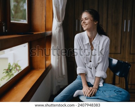 Woman sitting at home by a wooden window with a smile on a chair in homemade comfortable clothes and looking at the landscape, spring mood, women's day, rest on the weekend. Royalty-Free Stock Photo #2407948383