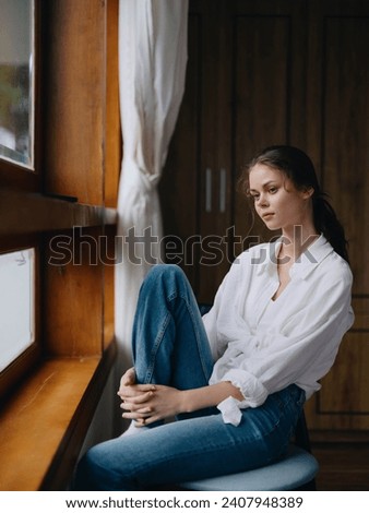 Woman sitting at home by a wooden window with a smile on a chair in homemade comfortable clothes and looking at the landscape, spring mood, women's day, rest on the weekend. Royalty-Free Stock Photo #2407948389