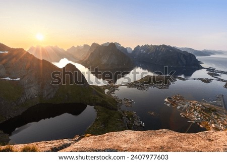 The midnight sun crests the rugged peaks at Reinebringen, shining over Lofoten Islands; its reflection shimmers across the fjord, illuminating the cozy village of Reine Royalty-Free Stock Photo #2407977603