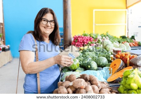  Joyful mature woman at a market, holding money and wallet, reflects a happy consumer having a great shopping experience on Consumer Day. Royalty-Free Stock Photo #2408002733