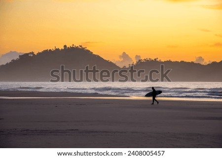 The picture shows the flat, sandy beach of Campeche on Santa Catarina Island at dawn. A surfer walks along the beach. In the background, you see Campeche Island and a golden sky.