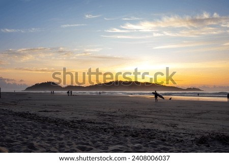 The picture shows the flat, sandy beach of Campeche on Santa Catarina Island at dawn. Surfers and early risers walk along the beach. In the background, you see Campeche Island and a golden sky.