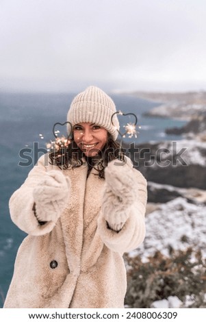 Outdoor winter portrait of happy smiling woman, light faux fur coat holding heart sparkler, posing against sea and snow background