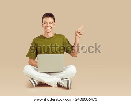 Portrait happy young man or teenage boy with laptop computer sitting on floor on beige studio background pointing index finger away showing information and inviting students to digital hackathon event