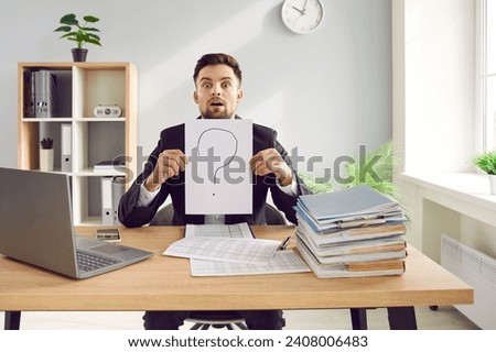Young accountant man employee sitting at office work desk with laptop and paper files and folders, holding sheet of paper with question mark and looking at camera with funny confused face expression