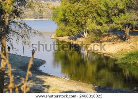 people fishing along the banks of Puddingstone Lake in a beautiful summer landscape with rippling water surrounded by lush green trees, plants and grass in San Dimas California USA Royalty-Free Stock Photo #2408018025