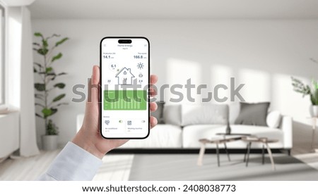 Hand holds phone displaying solar energy app in a cozy living room, effortlessly manage and monitor green power for sustainable living Royalty-Free Stock Photo #2408038773