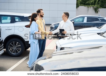 Car dealership employee communicates with couple of clients before test drive Royalty-Free Stock Photo #2408058033