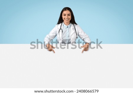 Smiling female doctor pointing downwards at blank white banner for text or graphics, with professional appearance and blue background, banner