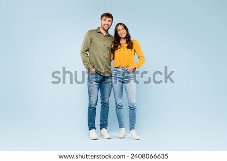Happy couple standing close in denim jeans and colorful shirts, sharing a comfortable moment together, all set against calm blue background, full length Royalty-Free Stock Photo #2408066635