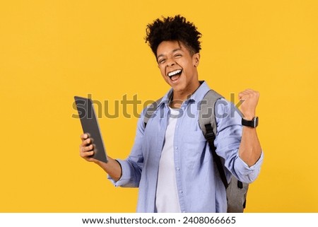 Victorious black male student holding tablet and fist-pumping in triumph, expressing sheer joy and achievement, all on yellow background Royalty-Free Stock Photo #2408066665